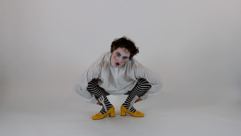 The performer is seen centre shot squated down, hands behind their ankles, with their feet in bright yellow box heels pointed in opposite directions, they wear striped tights and an oversized collarless white shirt, their hair is scruffy with one curl on their forehead, with rosy cheeks, dark eyes and red round lips they stare into the camera.