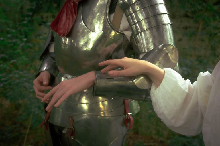 An editorial image with a dreamlike tone of a soft female hand reaching towards a knights arm, clad in shining armour.
