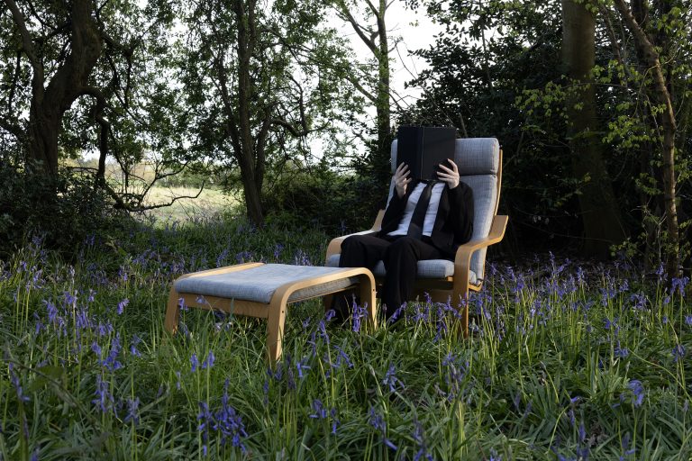 Photography Piece by Chloe Kirkland showing a person sitting on a chair reading a book in the middle of a bluebell field.