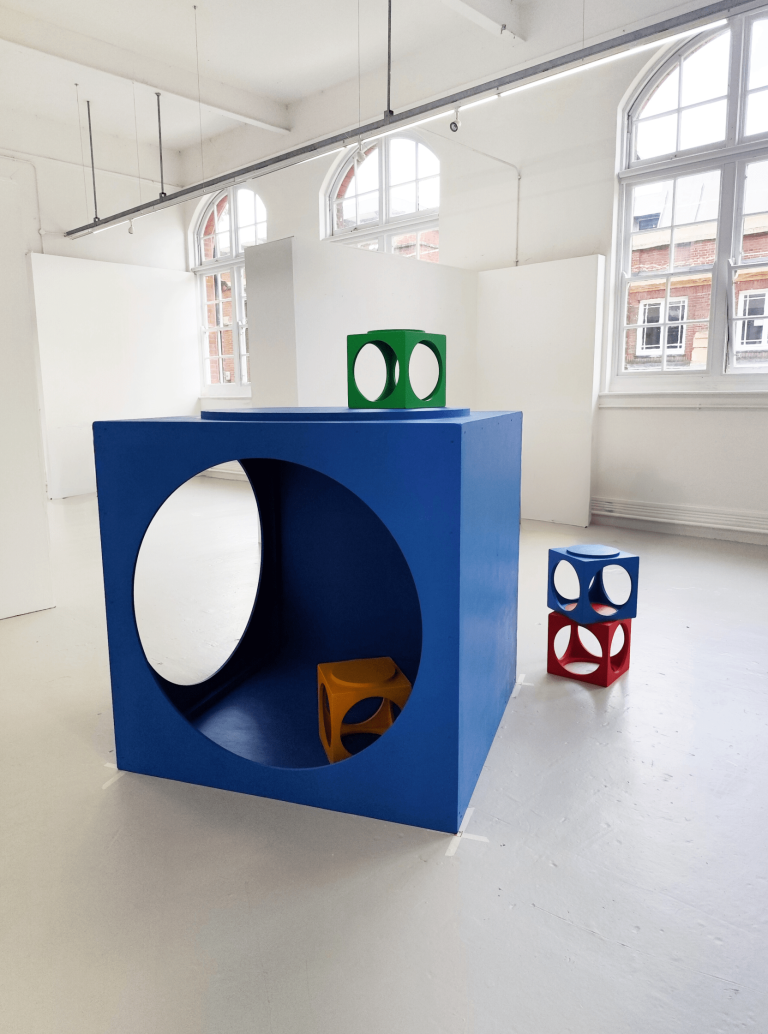 Fine Art work by Jess Haynes showing a bold blue MDF cube with circular openings; smaller blocks are arranged with one on top, one inside, and two outside.