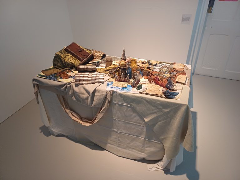 BA Fine Artwork by Toby Baker showing a table spread of various objects with a fantasy setting, the table is filled with trinkets, textures, scales and teeth.
