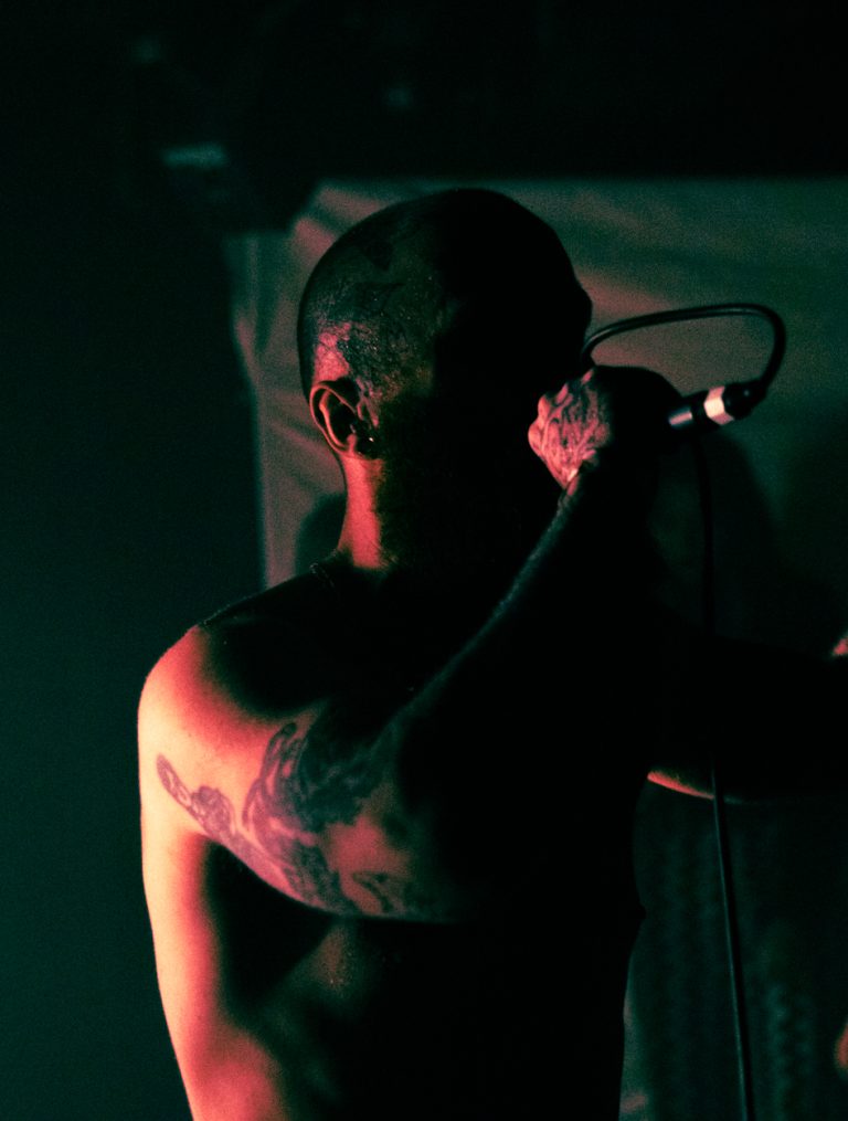 BA Photography work by Madeleine Townsend depicting a musician singing into a microphone. It is a low-light portrait, with strong shadows.