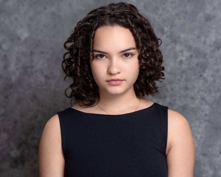 A person with short curly hair wearing a sleeveless black top stands against a gray textured background, looking directly at the camera with a neutral expression.