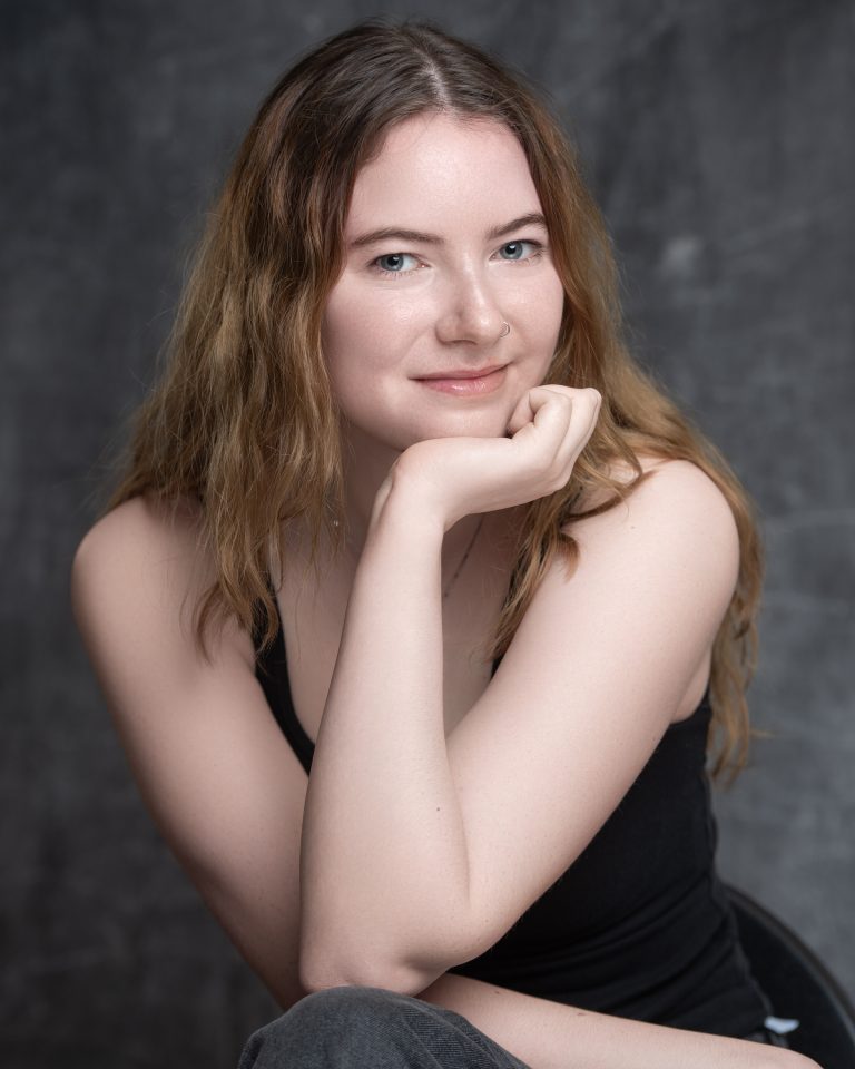 A person with long, wavy hair sits on a chair, resting their chin on their hand. They are looking at the camera and smiling against a grey background.