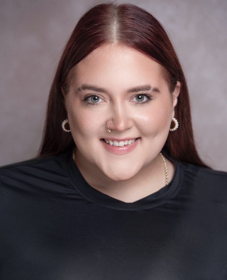 A person with long, straight, dark red hair parted down the middle, wearing hoop earrings and a nose ring, smiles warmly. They are dressed in a black top against a softly blurred background.