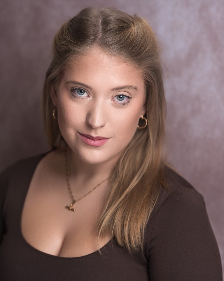 A person with long dark-blonde hair is looking directly at the camera and a slight smile. They are wearing a gold necklace and hoop earrings, and a brown coloured top and are against a grey background.