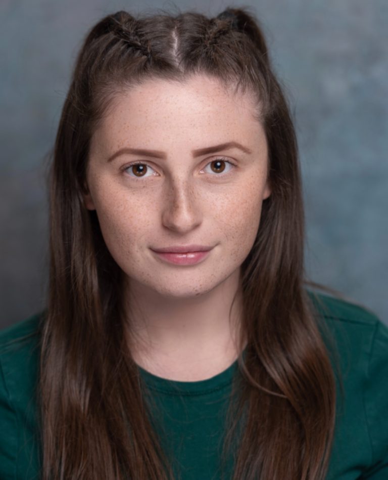 A person with long, straight brown hair styled in two braids on top and freckles across their face looks directly at the camera with a slight smile. They wear a dark green shirt with a neutral, textured background.