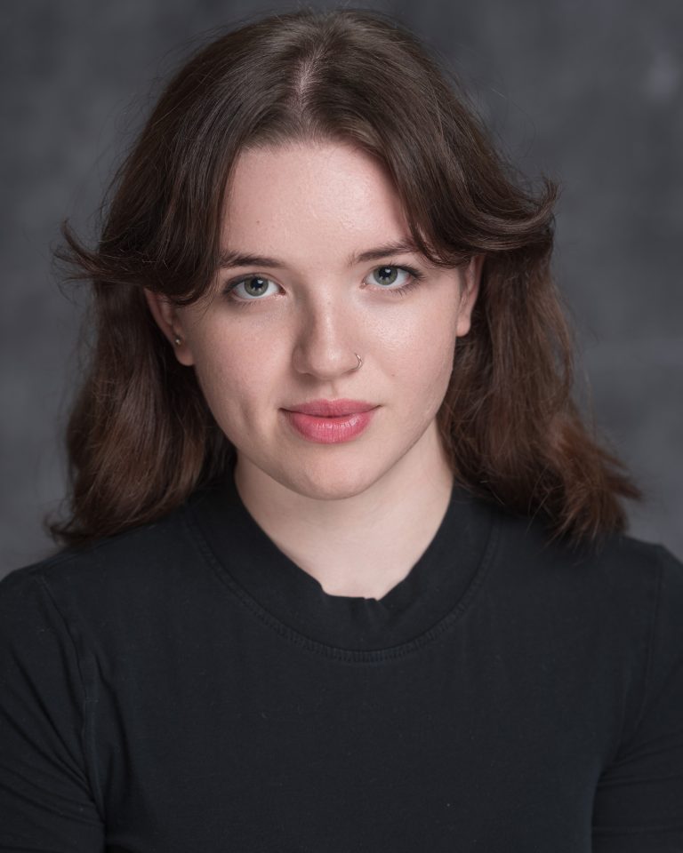A person with shoulder-length brown hair and a nose ring, wearing a black shirt, looks directly at the camera with a neutral expression against a dark, blurred background.
