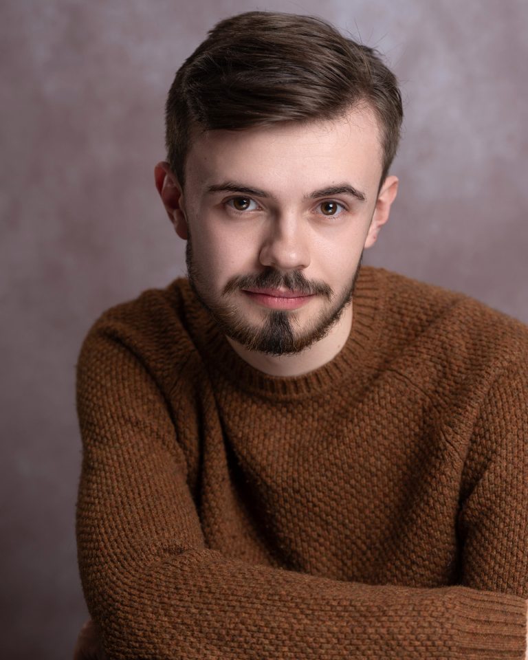 A person with short brown hair and a trimmed beard, wearing a brown knit sweater, sits with their arms crossed and looks into the camera against a muted brown background.