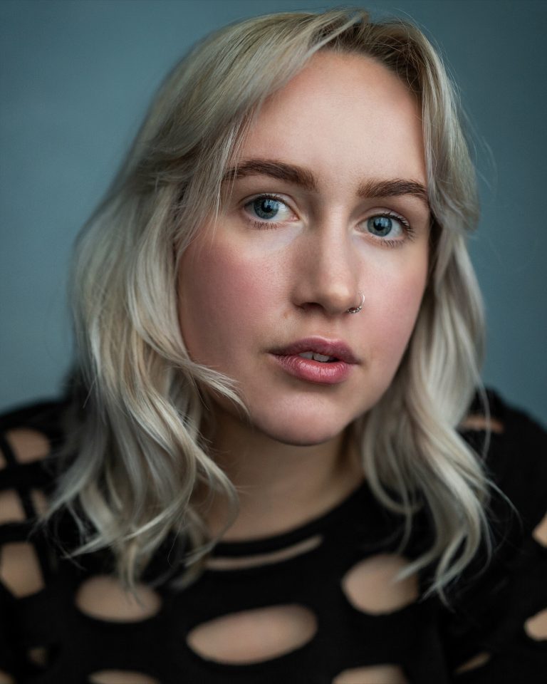 A person with light blonde hair and blue eyes looks at the camera. They are wearing a black top with circular cutouts and have a nose ring. The background is plain and blue-gray.