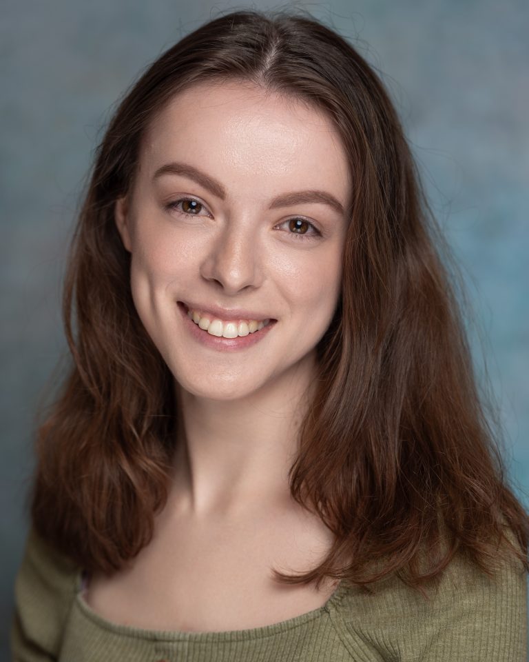A person with long brown hair smiles at the camera. They are wearing a green top and is posed against a soft blue and gray background.