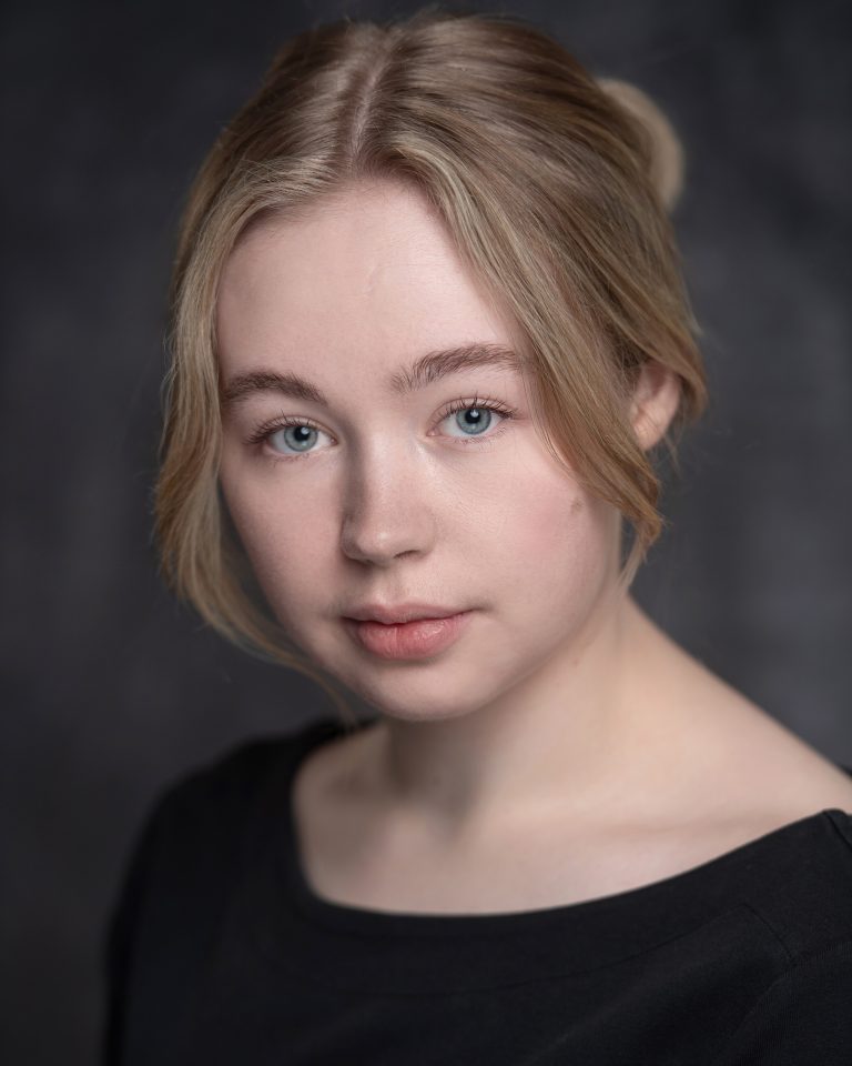 A person with blonde hair and blue eyes looks directly at the camera. They are wearing a black top against a neutral, dark gray background.