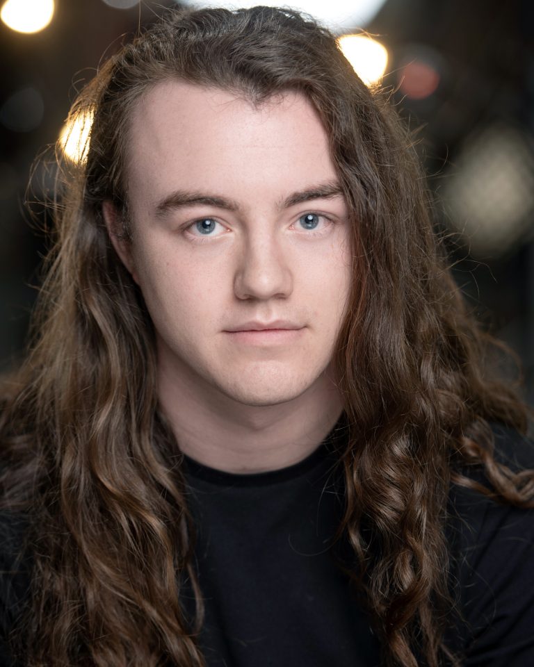 A young person with long, wavy brown hair and blue eyes looks at the camera, wearing a black shirt. The background is softly blurred with some lights visible.