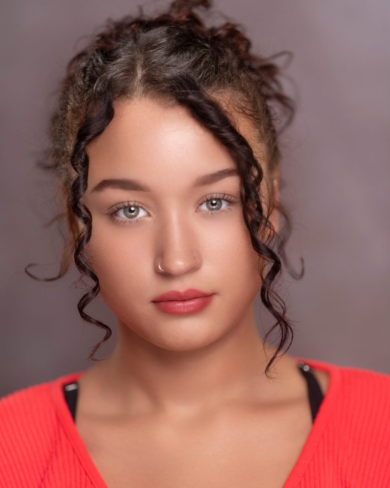 A person with curly brown hair, wearing a red top and a nose ring, looks directly at the camera with a neutral expression and a soft, blurred background.