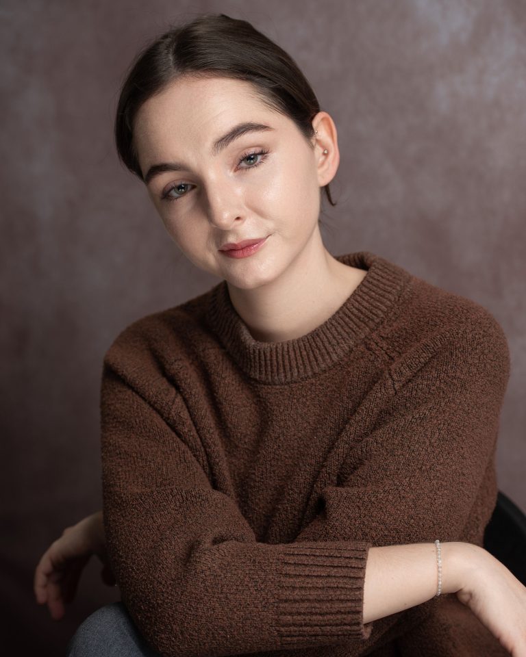 A person with brown hair, wearing a brown knit sweater, gazes softly at the camera against a neutral, softly blurred background.