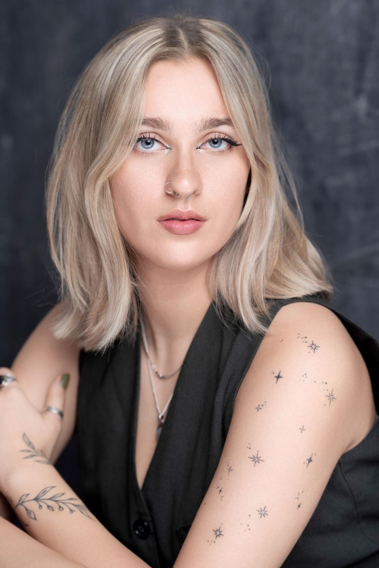A person with shoulder-length blonde hair and light blue eyes looks confidently at the camera. They have a nose ring, wear a sleeveless black top, and have tattoos of stars and leaves on their arms. The background is dark.