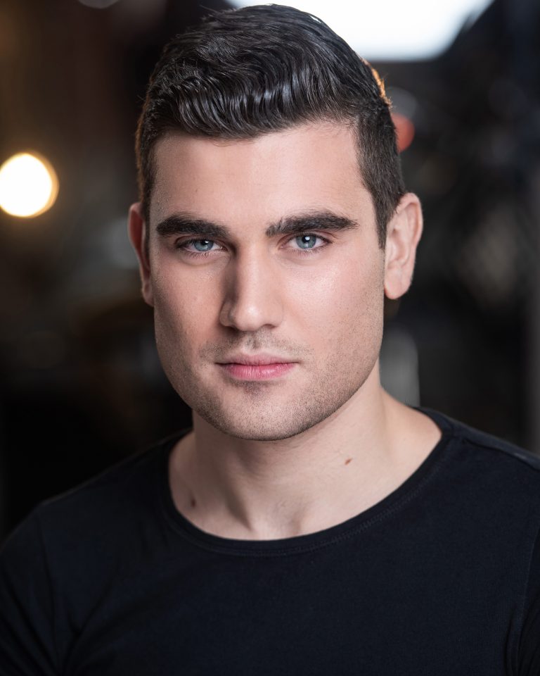 A person with short dark hair and light eyes wearing a black shirt, looking directly at the camera with a neutral expression, against a blurred dark background.