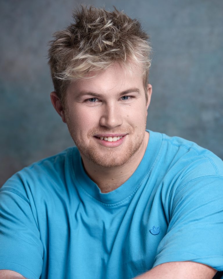 A person with messy blond hair and a short beard wearing a light gray ribbed sweater, looking directly at the camera with a neutral expression against a grey textured background.