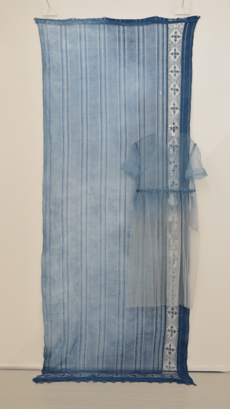 A see-through tulle dress is overlaid onto an indigo-dyed vintage curtain. The dress looks like it's blending into the wall, almost invisible.