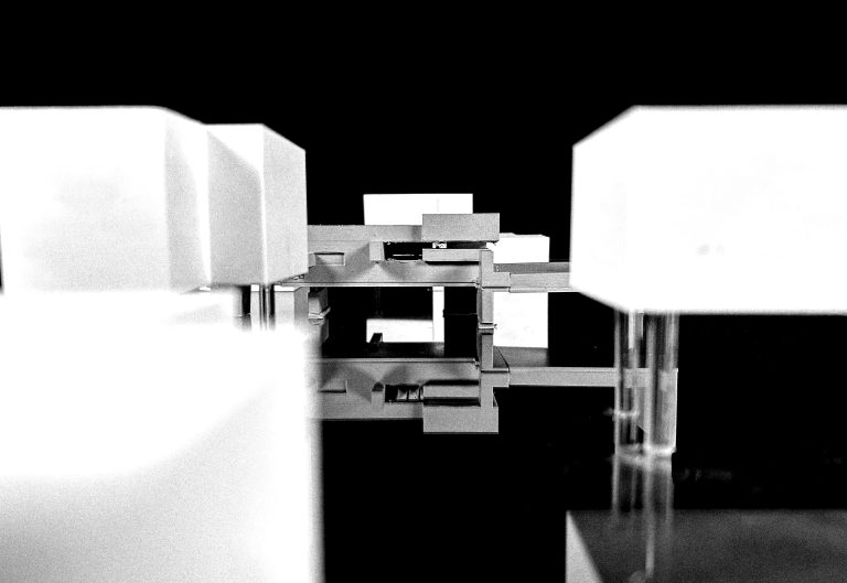 A floating arrangement of simple rectangular forms extends outward from a central structure. Suspended transparent supports create separation between volumes