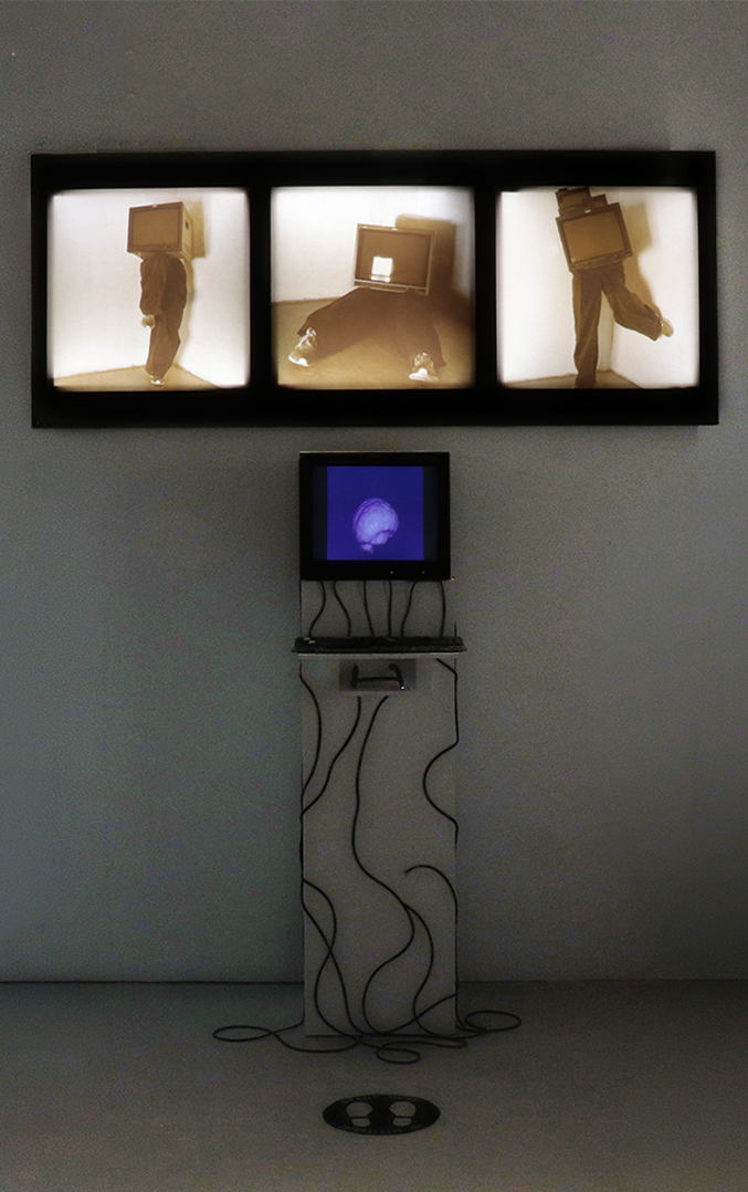 A gallery installation with three illuminated images above a small monitor showing a digital brain, with cables hanging below.