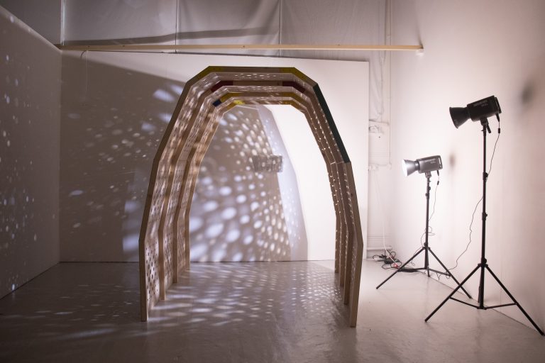 Installation artwork by Franka Matthes Thompson exhibits an arched tunnel of wood with a pattern of small holes, illuminated by spotlights, creating a pattern of light and shadows