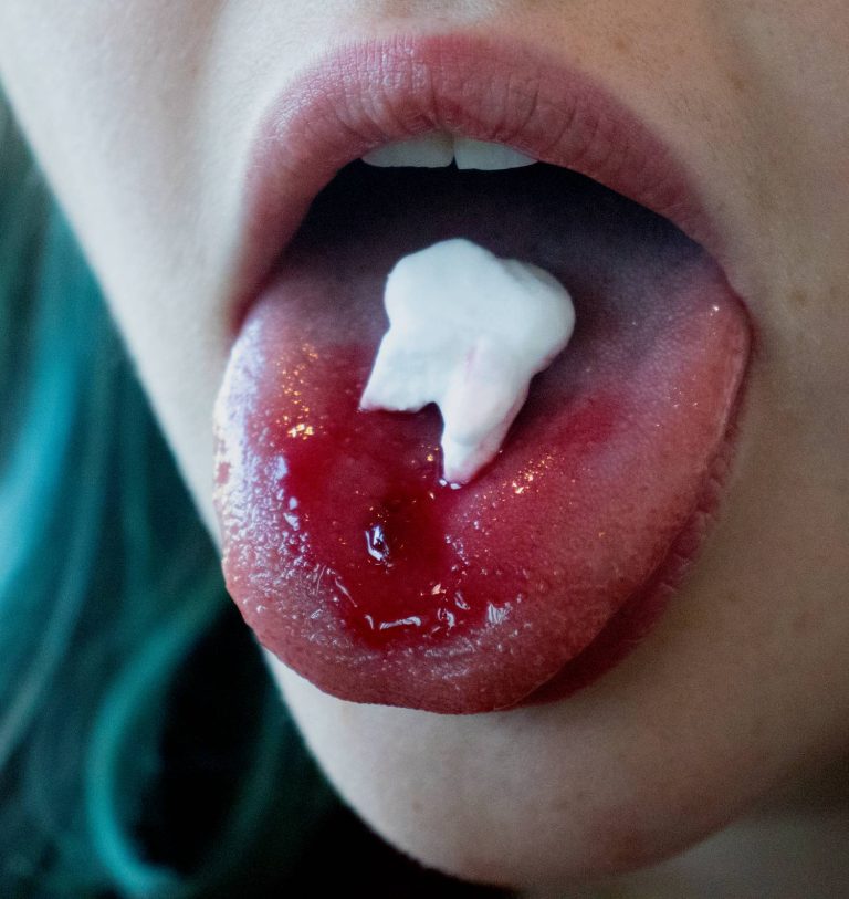 Fine Art work by Amy White documenting an eating performance exploring tooth loss phobia. The photograph depicts a tongue coated in strawberry syrup, cushioning a peppermint carved into a tooth.