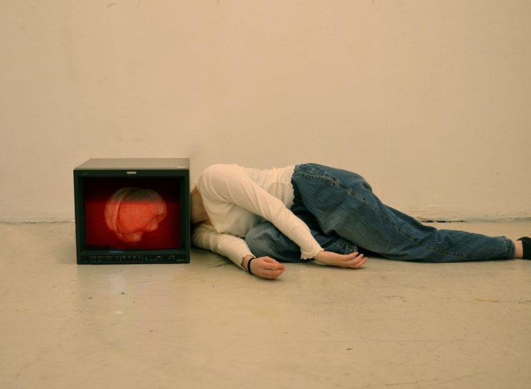 A person lies on the floor beside a monitor displaying a red, distorted brain-like image.