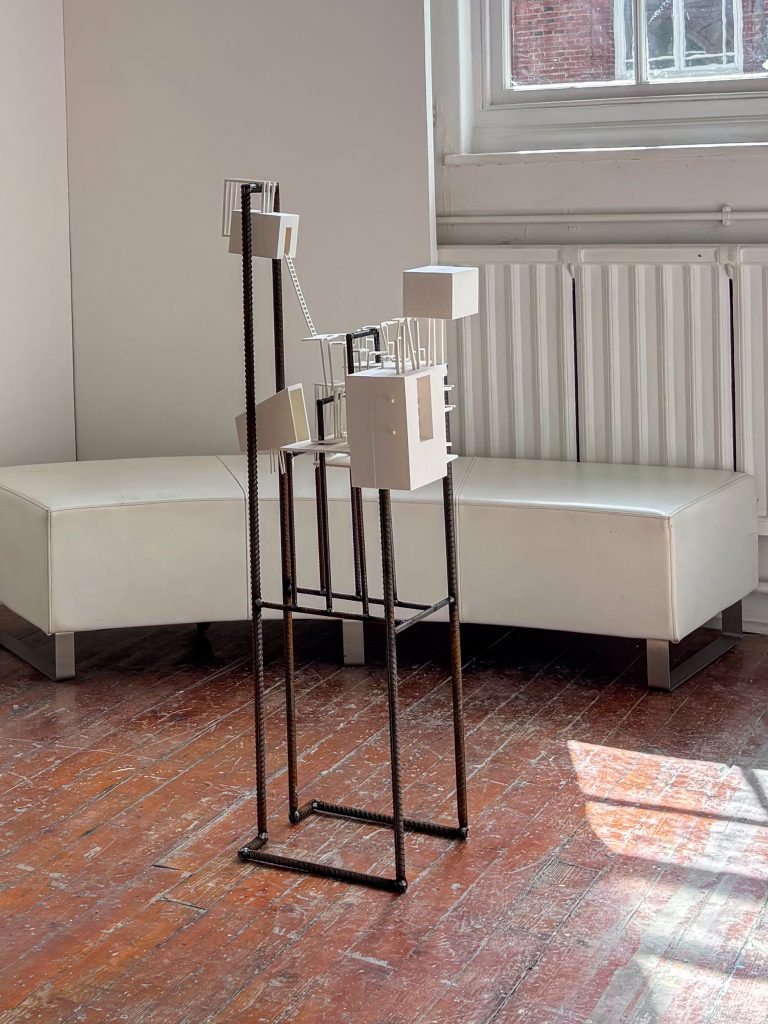 A geometric rebar sculpture containing suspended white architectural forms stands on a wooden floor within a gallery space, interacting with light, shadow, and the framing of a large sunlit window.