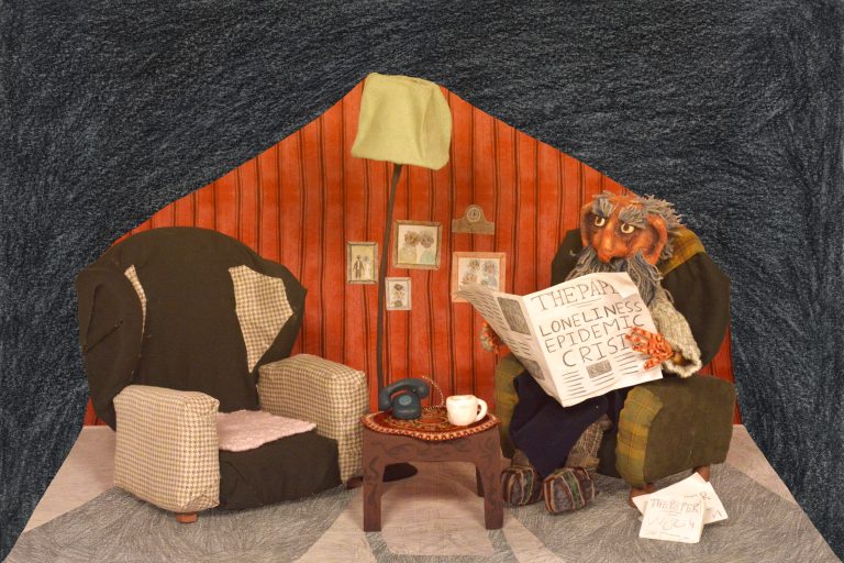 A lounge featuring red vintage wallpaper, two green armchairs, wooden coffee table and green standing lamp. An elderly gentleman sits in one chair, reading a newspaper, headlined ‘LONELINESS EPIDEMIC CRISIS’.