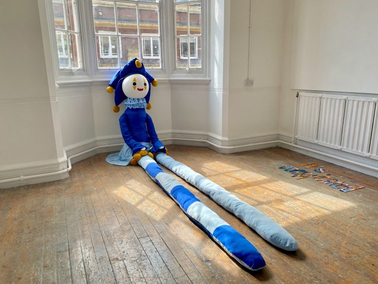 A 1.5m tall, clown-like doll sat in a bay window, it has 3m long legs stretched out in front of it.