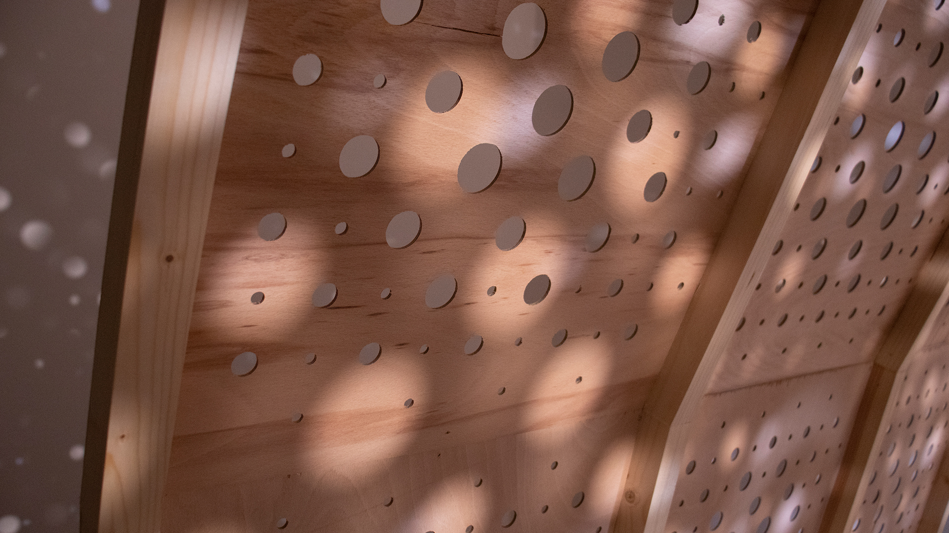 Video of an installation artwork featuring arched tunnel covered in a pattern of small circular holes illuminated by spotlights, showing details of light and shadows, holes in plywood, and painted beams.